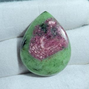 Op de afbeelding: Een druppelvormige edelsteen met een levendige kleurencombinatie. De steen heeft een lichtgroene basis met een opvallend, getextureerd, magenta-kleurig centrum. Donkerdere zwarte en groene insluitsels zijn verspreid over de steen, wat diepte en detail toevoegt.