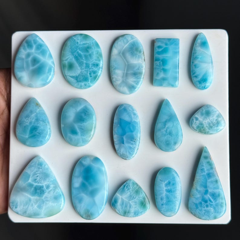 Puede incluir: Una colecci&oacute;n de catorce cabujones de larimar pulidos dispuestos sobre un tablero blanco. Las piedras presentan formas variadas, incluyendo gotas, &oacute;valos, rect&aacute;ngulos y tri&aacute;ngulos, todas con un color azul cielo caracter&iacute;stico y patrones blancos.