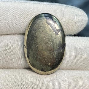 May include: Oval-shaped pyrite gemstone with a metallic luster. The stone has a mix of gold, bronze, and dark gray colors, with visible natural patterns and textures. The polished surface reflects light, highlighting its unique mineral composition.