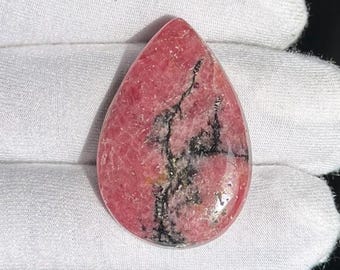 May include: A teardrop-shaped, polished gemstone with a vibrant pink hue. The stone features striking black veining and a glossy surface. The gemstone is set against a white background, highlighting its color and texture.