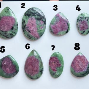 Puede incluir: Ocho piedras preciosas de zoisita pulida con inclusiones de rubí, dispuestas en dos filas sobre una superficie blanca. Cada piedra está numerada del uno al ocho, mostrando una mezcla de patrones minerales en rosa, verde y negro.