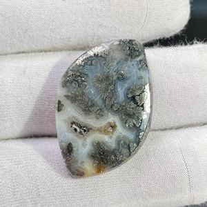 Puede incluir: Una piedra preciosa pulida de forma irregular, con un patrón único. La piedra presenta una mezcla de tonos grises, plateados y blancos, con intrincadas formaciones en forma de árbol. El aspecto general es liso y reflectante, lo que sugiere un acabado de alta calidad.