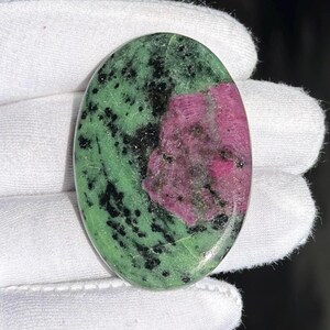 Puede incluir: Gema de forma ovalada con una vibrante mezcla de colores. La piedra tiene una base verde con motas negras y una sección prominente de color rosa intenso. La superficie pulida refleja la luz, resaltando los patrones y texturas naturales de la piedra.