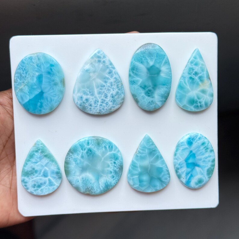 May include: A collection of eight polished larimar cabochons arranged on a white rectangular display card. The stones feature various shapes including circles, ovals, and teardrops, all displaying a vibrant sky-blue colour with white patterns.