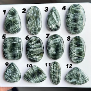 Puede incluir: Una colección de doce cabujones de serafinita pulidos dispuestos sobre un tablero blanco. Cada piedra presenta una base de color verde oscuro con inclusiones plateadas y plumosas. Las piedras están numeradas del uno al doce.
