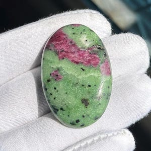 Op de afbeelding: Ovaalvormige edelsteen met een levendige kleurenmix. De steen toont een opvallende combinatie van dieprode en groene tinten, met zwarte spikkels over het oppervlak. De gepolijste steen wordt in een witte handschoen gehouden. Ongeveer 4 cm lang.