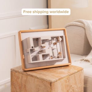 Könnte beinhalten: Ein gerahmtes Architekturmodell mit hellem Holzrahmen. Das Modell ist eine detaillierte, dreidimensionale Darstellung eines Innenraums, mit beigen Wänden und braunen Akzenten. Der Rahmen ruht auf einer marmorierten Oberfläche.