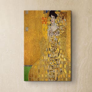 May include: A framed canvas print of a Gustav Klimt painting, featuring a woman in a gold dress with intricate patterns. The artwork is displayed on a beige wall above a round table and chairs, creating a gallery-like atmosphere.