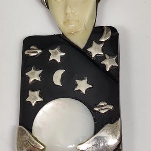 May include: A celestial-themed brooch featuring a cream-colored face and silver accents. The black background is adorned with silver stars, moons, and UFOs. A large, white, round pearl is held by silver hands. The brooch has a silver border.
