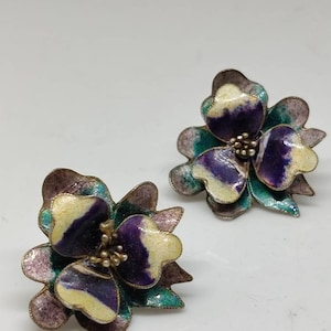 May include: A pair of vintage flower-shaped earrings with purple, white and green enamel. The earrings are made of metal and have a delicate design.