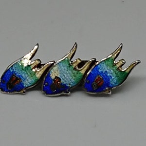 May include: A silver brooch with three enamel fish in blue, green, and white. The fish have gold accents and red eyes.