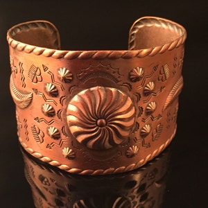 May include: A copper cuff bracelet featuring a swirling design in the centre and geometric patterns around the edges.