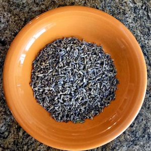 May include: An overhead shot of an orange ceramic bowl filled with dried lavender buds. The lavender buds are a mix of purple and gray, and the bowl is set against a speckled, dark brown surface.