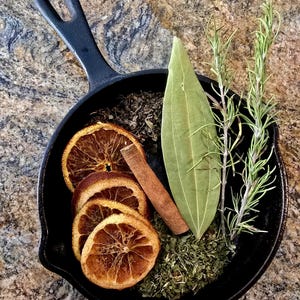May include: A black cast iron skillet filled with dried orange slices, a cinnamon stick, a bay leaf, rosemary sprigs, and dried herbs. The text "Abundant Alchemy" is above the skillet.