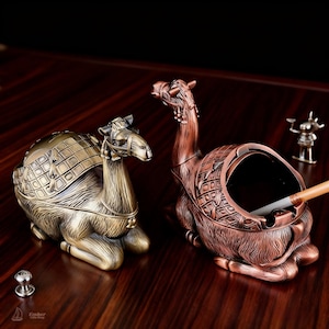 Pode incluir: Dois cinzeiros decorativos em forma de camelo em tons de bronze e cobre. Os cinzeiros apresentam designs detalhados e estão colocados sobre uma superfície de madeira escura. Um tem tampa, o outro contém um cigarro aceso.