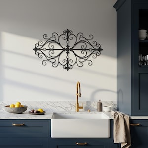 Könnte beinhalten: Schwarze Metall-Wanddekoration mit einem verzierten, symmetrischen Design. Das dekorative Stück zeigt wirbelnde Muster und eine zentrale Verzierung, montiert über einem Küchenbecken mit einer goldenen Armatur. Die Küche hat marineblaue Schränke und eine weiße Arbeitsplatte.