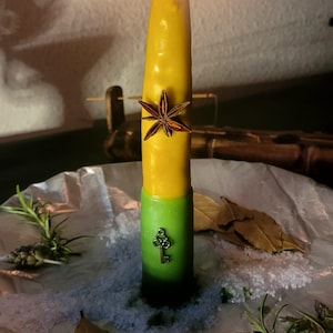 Può includere: Una candela affusolata accesa con la metà superiore gialla e quella inferiore verde. Un anice stellato marrone è incastonato nella sezione gialla e una piccola chiave di metallo in quella verde. La candela poggia su una base di sale grosso e rosmarino.