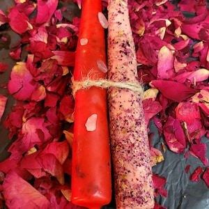 Puede incluir: Dos velas cónicas atadas con hilo, una roja brillante y otra rosa claro. La vela roja tiene pequeños cristales transparentes adheridos. La vela rosa está cubierta de pétalos de rosa secos. Las velas están rodeadas de pétalos de rosa rojos esparcidos.