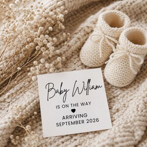 May include: A baby announcement card with the name "Baby William" and the text "Is on the way, arriving September 2026." Two cream-colored knitted baby booties are next to the card, on a cream-colored knitted blanket.