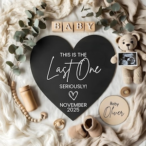 Pode incluir: Uma placa preta em forma de coração com o texto branco "THIS IS THE Last One SERIOUSLY!" e "NOVEMBER 2025". A imagem inclui blocos de madeira "BABY", um ursinho de pelúcia, sapatos de bebê e outros itens para bebês.