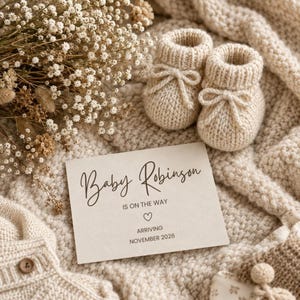 Puede incluir: Un par de patucos de punto beige con lazos, una tarjeta de anuncio color crema que dice Baby Robinson IS ON THE WAY con un corazón y ARRIVING NOVEMBER 2026, y un ramo de flores blancas secas sobre un tejido de punto color crema.