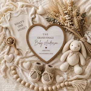 May include: A cream-colored baby bodysuit with the text WHAT'S ONE MORE?, a heart-shaped letter board reading THE GRAND FINALE Baby Andreson ARRIVING NOVEMBER 2026, a plush rabbit, wooden rattle, knit booties, and a wooden bead garland on a lace fabric.