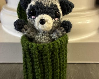May include: A handmade crochet raccoon toy in a green container. The raccoon has a gray and white body, black ears, and large black eyes. The container is a dark green color and has a ribbed texture.
