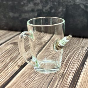 Puede incluir: Una taza de vidrio transparente con asa, con una bujía adjunta en el lateral. La taza es transparente. La bujía tiene una base metálica y un aislante cerámico blanco. La taza está sobre una superficie de madera.