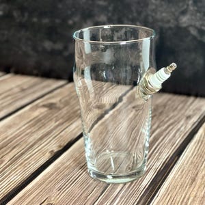 Könnte beinhalten: Ein klares Glas mit einer Zündkerze an der Seite. Das Glas ist zylindrisch und steht auf einer Holzoberfläche. Die Zündkerze ist silber und weiß.
