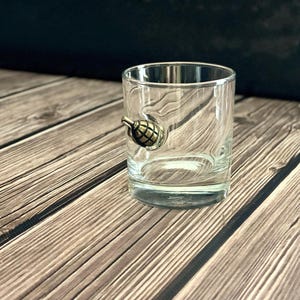 以下が含まれることがあります： 側面にブロンズ色の装飾的な手榴弾が付いた、透明で短いタンブラーグラス。 グラスは、暗い背景のある木製の表面に置かれています。
