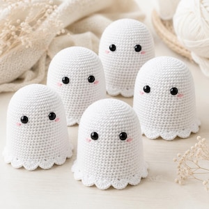 May include: Five white crocheted ghost figures with rounded tops and scalloped bottom edges. Each ghost features two small black circular eyes and two tiny pink embroidered blush marks on the cheeks, crafted from white yarn in a tight stitch.