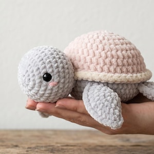 May include: A small, handmade crochet turtle plush toy featuring a soft, light gray head and flippers, a pale pink shell with a cream-colored border, and two round, black plastic eyes with small pink stitched cheeks on the gray yarn head.