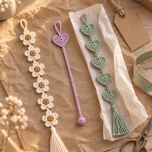 May include: Three handmade crochet bookmarks. One features a vertical chain of six white flowers with yellow centres and a cream tassel. One is a single lavender heart on a long cord with a small ball end. One has five sage green hearts and a tassel.