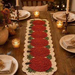 May include: A rectangular crochet table runner featuring six circular orange pumpkin motifs with dark brown stems and green leaves, bordered by a scalloped cream-colored edge. The textile piece is centered on a wooden table surface.