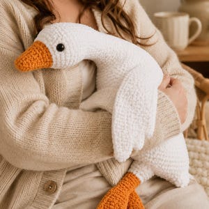May include: A white crocheted goose plush toy with a textured yarn finish, featuring a bright orange beak and matching orange webbed feet. The stuffed animal has a long neck, a rounded body, and a single black plastic eye visible on the side.