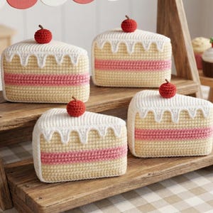 May include: Crocheted cake slices with white frosting, a pink layer, and a red cherry on top. The slices are displayed on a wooden stand with other crocheted desserts, including cupcakes and cookies. The scene evokes a playful bakery theme.