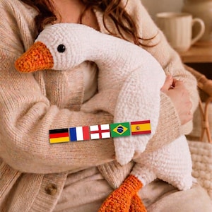 May include: A white crocheted goose plush toy with a textured, soft yarn body, a bright orange beak, and orange webbed feet. The goose features small black bead eyes and is decorated with a horizontal row of five small rectangular national flags.