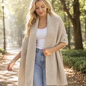 May include: A beige open-front knit cardigan featuring a textured stitch pattern, long sleeves, and a ribbed hem. The garment is worn over a plain white tank top and light blue denim jeans, showcasing a relaxed fit and a neutral color palette.