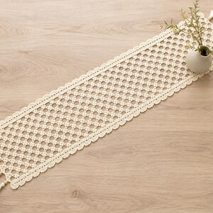 Puede incluir: Un camino de mesa rectangular de ganchillo color crema con un patrón calado circular repetitivo y un borde festoneado. El camino está rematado con pequeñas cuentas de madera y borlas colgantes en ambos extremos, junto a un pequeño jarrón blanco.