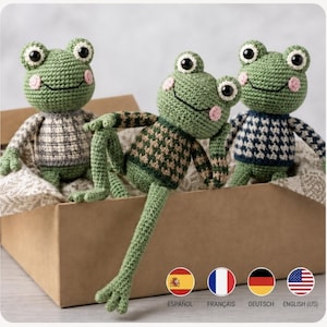 Könnte beinhalten: Drei handgefertigte Häkel-Froschpuppen mit grünen Körpern, rosa Wangen und großen schwarzen Augen. Jeder Frosch trägt einen anders gemusterten Pullover: einen mit grauem Karomuster, einen mit grün-braunem Schachbrettmuster und einen mit blau-weißem Hahnentrittmuster.