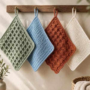 May include: Four square crochet dishcloths hanging from a wooden rack. The cloths are sage green, light blue, burnt orange, and cream, each featuring a distinct textured stitch pattern and a small loop at the top corner for convenient hanging.