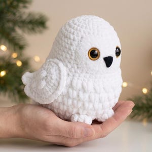 May include: A white crocheted owl plush toy featuring a rounded body, textured yarn stitches, large amber-colored plastic eyes with black pupils, and a small black triangular beak. The owl has detailed wing patterns and small feet.
