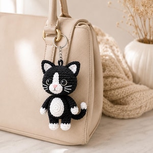May include: A black and white crocheted cat keychain featuring a small, stuffed feline figure with a white belly, white paws, and white inner ears. The cat has black plastic eyes, a pink stitched nose, and white whiskers attached to a metal ring.