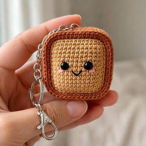 May include: A square, crochet-style protective case for wireless earbuds, featuring a light tan center and a darker brown border. The front displays a stitched face with black bead eyes, a small smile, and pink blush marks, attached to a silver keychain.