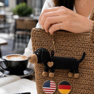 Puede incluir: Un bolso de mano tejido en color marrón con un llavero de perro salchicha tejido a ganchillo en negro y beige con un pequeño corazón, y dos pines circulares con las banderas de Estados Unidos y Alemania en la parte frontal.