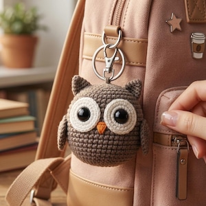 Pode incluir: Um porta-chaves de coruja em croché castanho com olhos pretos grandes, manchas brancas à volta dos olhos e um pequeno bico cor de laranja, preso a um mosquetão e argola de metal prateado, pendurado numa mochila de lona castanha clara com alfinetes.