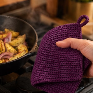 May include: A deep purple, square-shaped, hand-crocheted potholder featuring a small, integrated loop at the top corner for hanging. The fabric has a dense, textured stitch pattern, providing a thick layer for handling hot kitchen cookware.