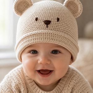 Op de afbeelding: Een beige gehaakte muts met twee kleine ronde berenoortjes aan de bovenkant en een eenvoudig geborduurd berengezicht met twee ogen en een neus. De muts is gemaakt met een dicht, uniform steekpatroon en heeft een omgeslagen rand aan de basis.
