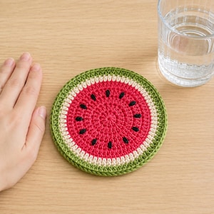 May include: A round, crocheted coaster designed to look like a slice of watermelon. The coaster features a bright red center with small black embroidered seeds, a thin white ring, and a thick green outer border made of yarn.