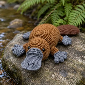 May include: A handmade crochet platypus toy featuring a brown body, a dark brown tail, and a textured grey bill and webbed feet. The plush animal has small black bead eyes and is crafted with visible yarn stitches, showcasing a detailed design.